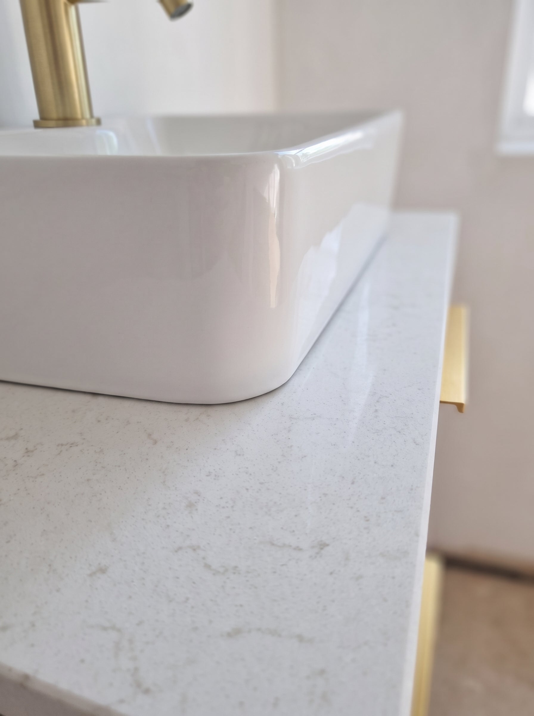 Close-up of a white rectangular vessel sink with a gold faucet, set on a white speckled countertop with a gold cabinet pull handle.