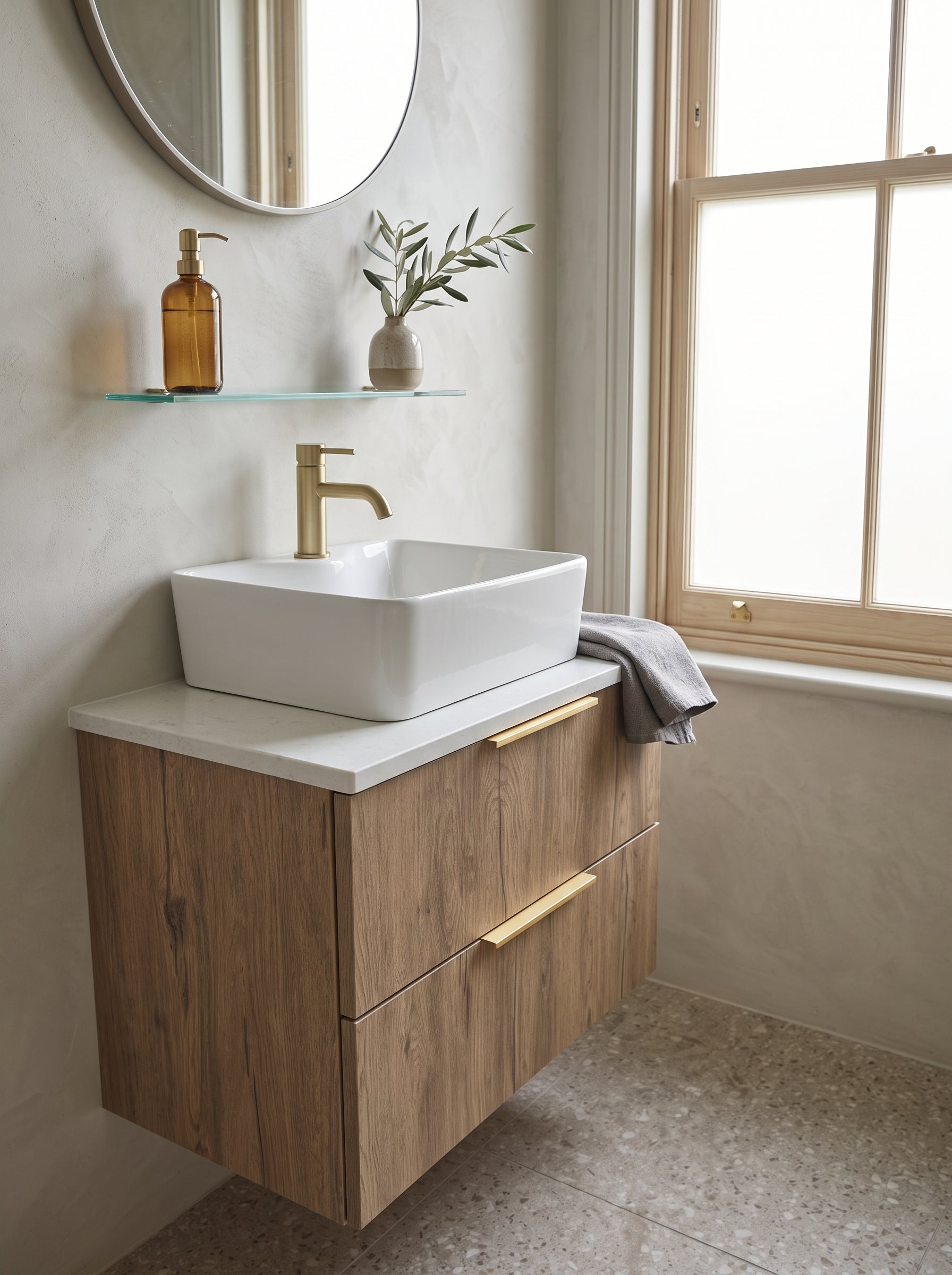 A modern floating bathroom vanity with a light oak wood grain cabinet, white countertop, and a white rectangular vessel sink. The vanity is styled in a bright bathroom with a gold faucet, a glass shelf with toiletries, and a window.