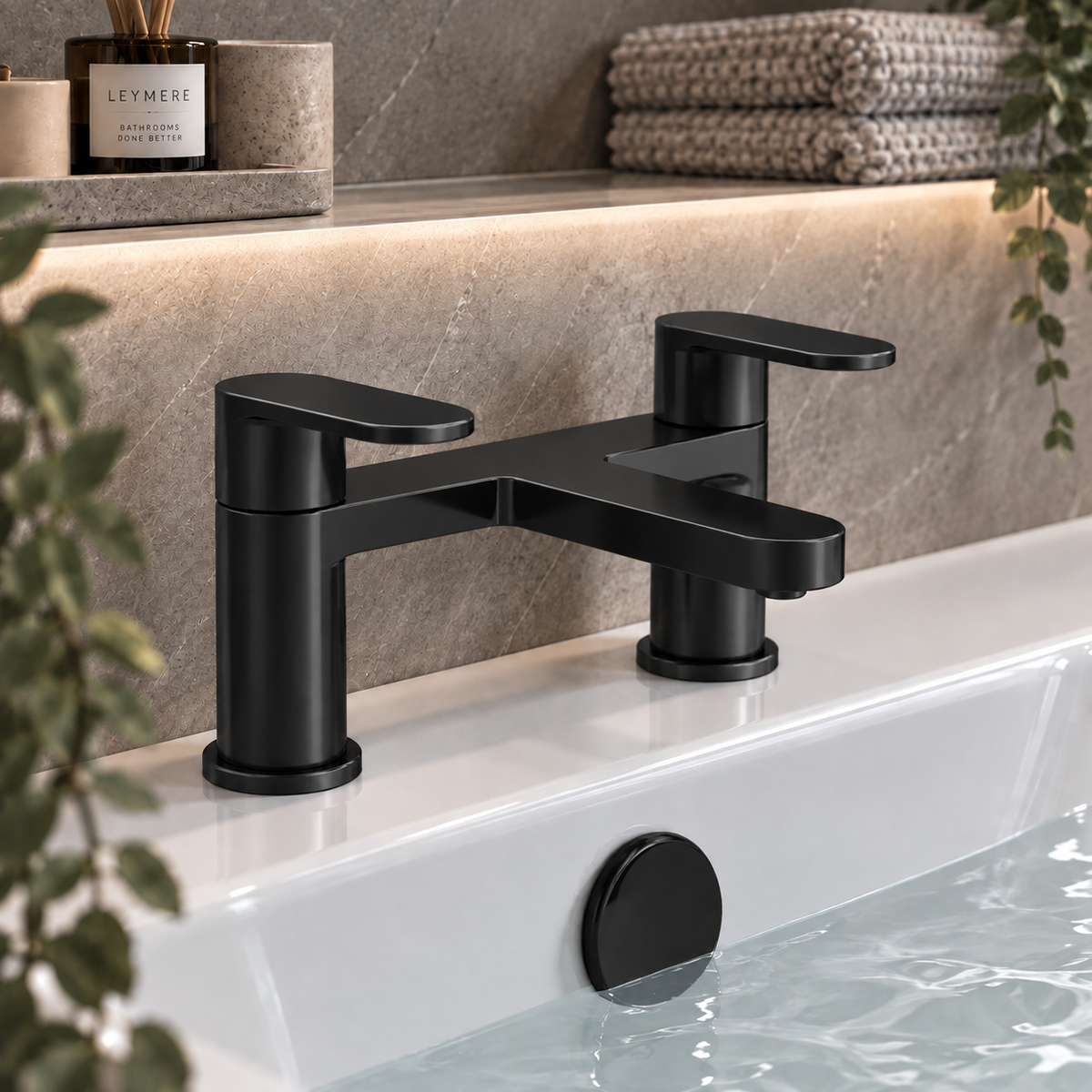 A matte black two-handle bathroom faucet mounted on a white sink, with a stone backsplash, folded towels, and greenery in the background.