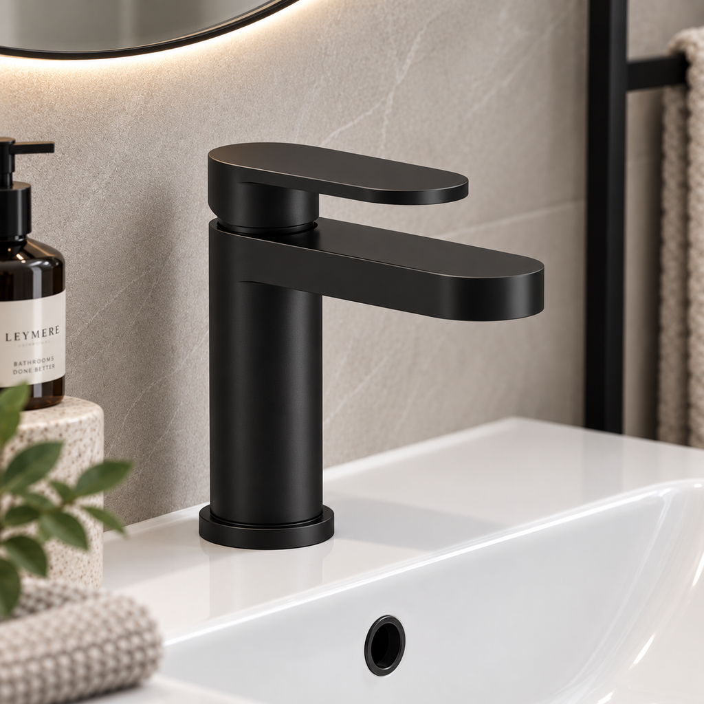 Matte black single-handle bathroom faucet installed on a white sink, with a tiled wall, mirror edge, soap dispenser, and towel in the background.