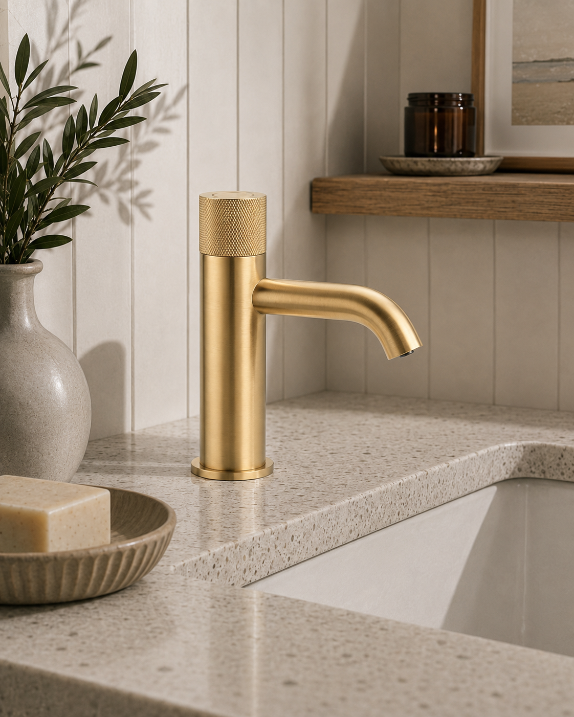 Gold faucet on a bathroom counter with a vase, soap, and plant in the background.