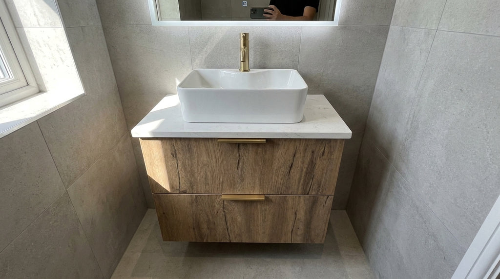 A modern floating bathroom vanity with a white rectangular vessel sink, a gold faucet, and a wood-grain cabinet, set against gray tiled walls.