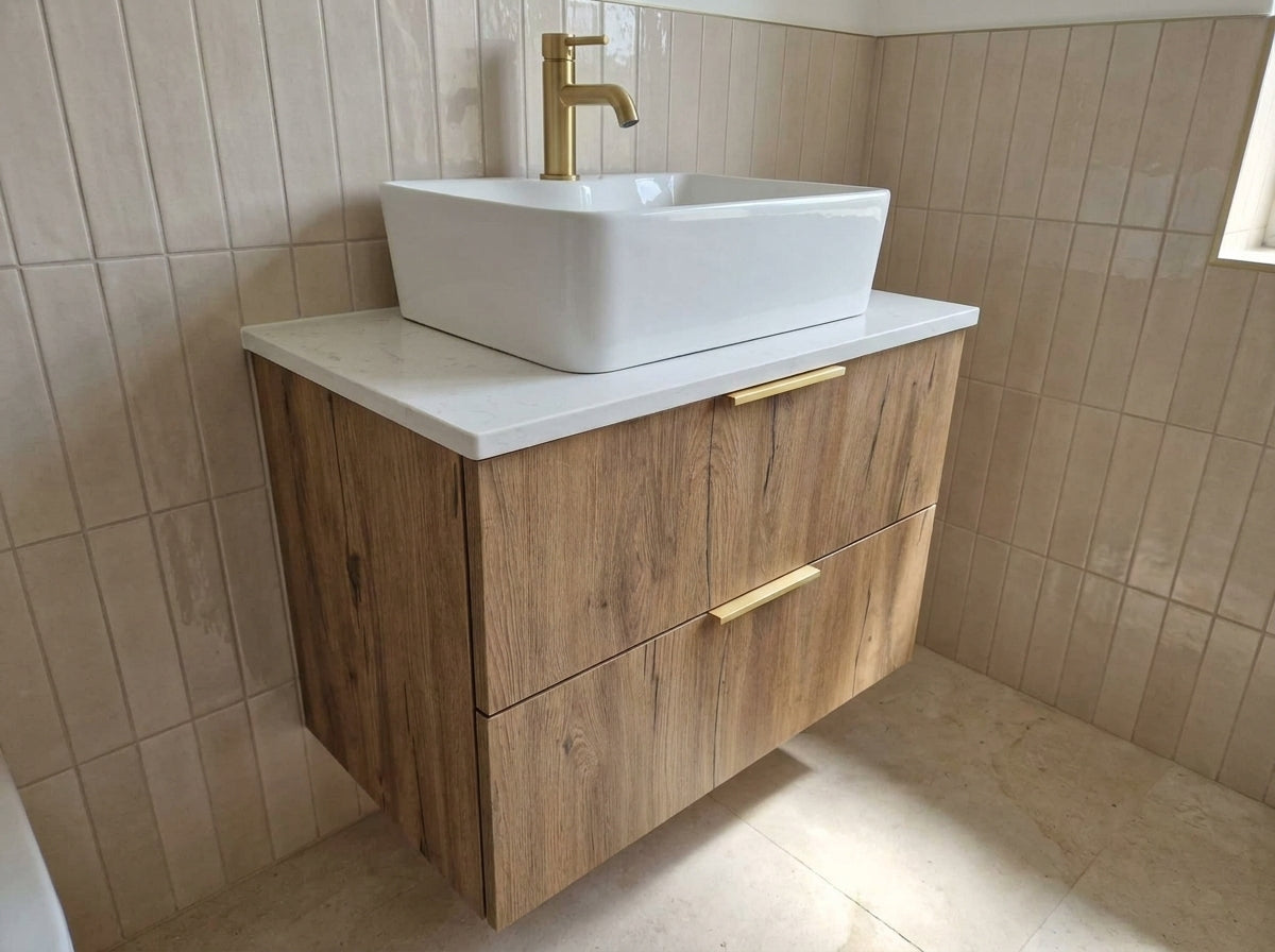 A modern floating bathroom vanity with a light oak wood cabinet, white countertop, white rectangular vessel sink, and a gold faucet, set against beige tiled walls.